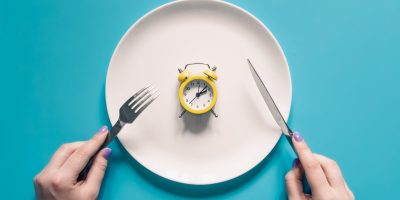 Hands holding knife and fork above alarm clock on a plate on blue background. Intermittent fasting, Ketogenic dieting, weight loss, meal plan, and healthy food concept.